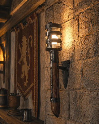 Realistic medieval-style wall torch light mounted on a textured stone wall inside a castle-inspired room. Handmade black wrought iron and wooden torch with a warm LED bulb glowing softly, surrounded by rustic decor and a cozy tavern atmosphere.